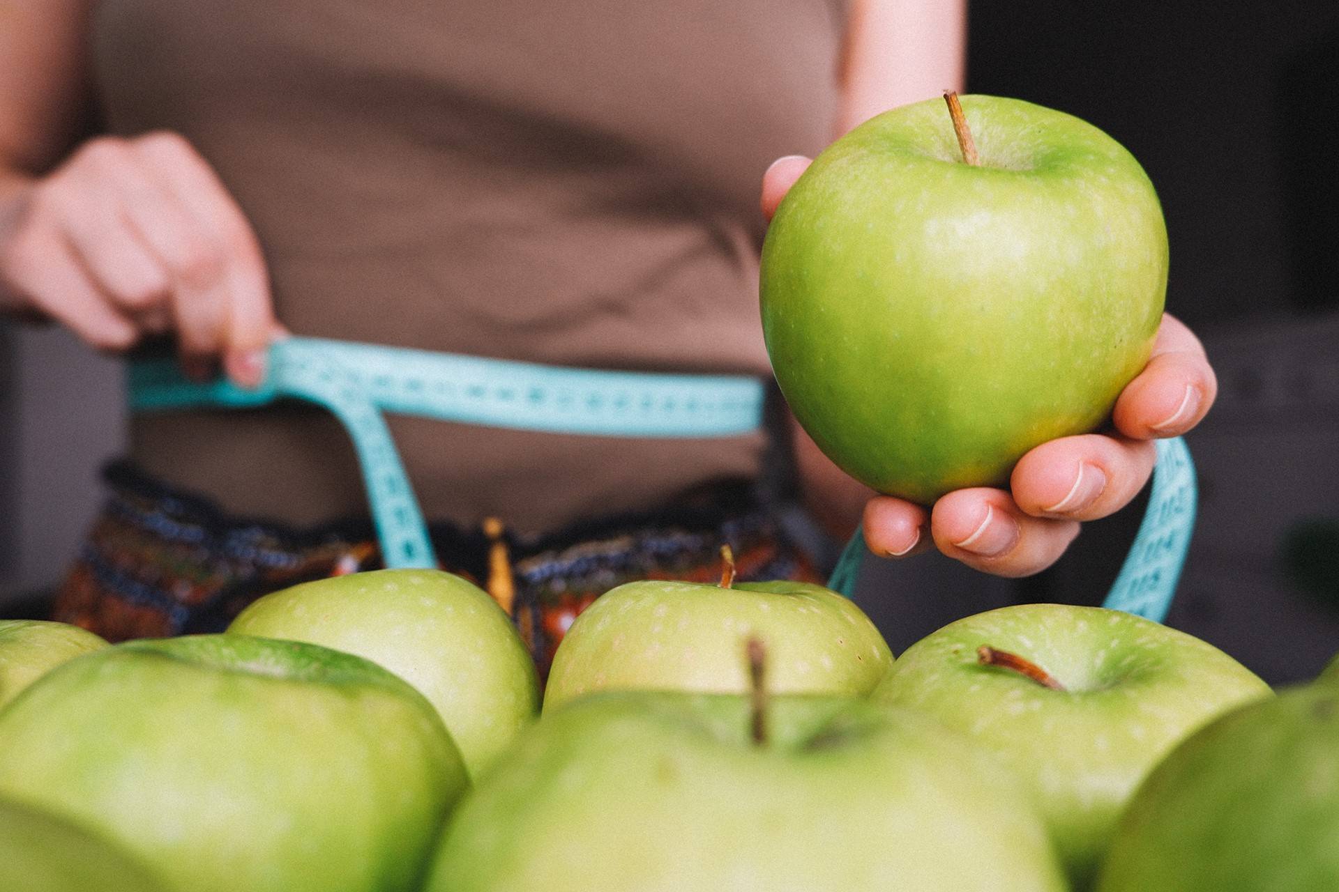 Жінка тримає зелене яблуко та вимірює талію — здорове харчування і контроль ваги для підтримки травної системи.