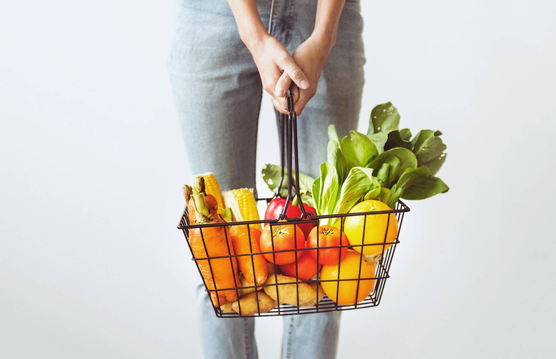 Жінка тримає в руках металевий кошик, наповнений свіжими овочами та фруктами — морквою, помідорами, картоплею, салатом, кукурудзою та апельсинами.