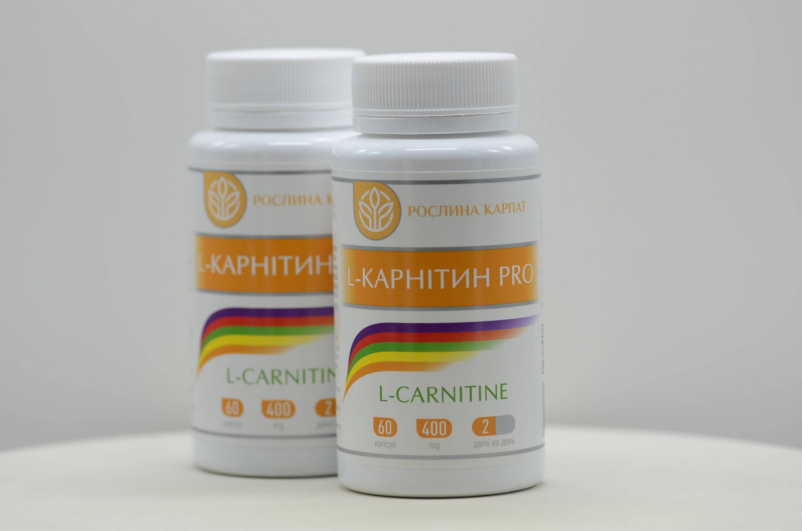 Дві баночки харчової добавки L-карнітин PRO від «Рослина Карпат» із яскравою помаранчевою етикеткою та кольоровою смугою, розташовані на світлому фоні.